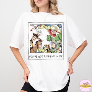 以下が含まれることがあります： 白のTシャツ。映画「トイ・ストーリー」のキャラクター、ウッディとバズ・ライトイヤーを含むイラストがフレームに入っています。画像には、映画の他のキャラクターも含まれています。「YOU'VE GOT A FRIEND IN ME」の文字が下に印刷されています。