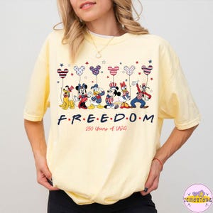 May include: A light yellow t-shirt featuring a graphic of Disney characters holding heart-shaped balloons in red, white, and blue. The word "FREEDOM" is printed below the characters, with "250 Years of USA" underneath.
