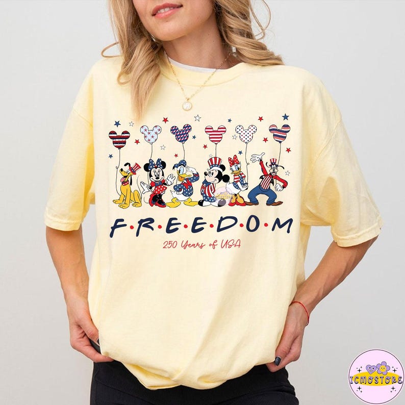 May include: A light yellow t-shirt featuring a graphic of Disney characters holding heart-shaped balloons in red, white, and blue. The word "FREEDOM" is printed below the characters, with "250 Years of USA" underneath.
