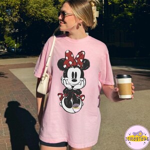 May include: A light pink t-shirt featuring a Minnie Mouse graphic. The cartoon character is depicted in black, white, and red, with a polka-dot bow. The shirt is a casual, oversized style.