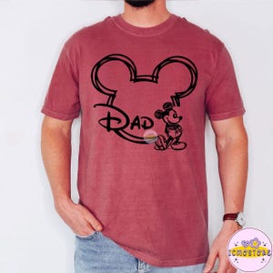 May include: A maroon t-shirt featuring a black outline of Mickey Mouse ears with the word "Dad" in a stylized font. A small Mickey Mouse graphic is on the right side. The shirt is a solid color and has short sleeves.