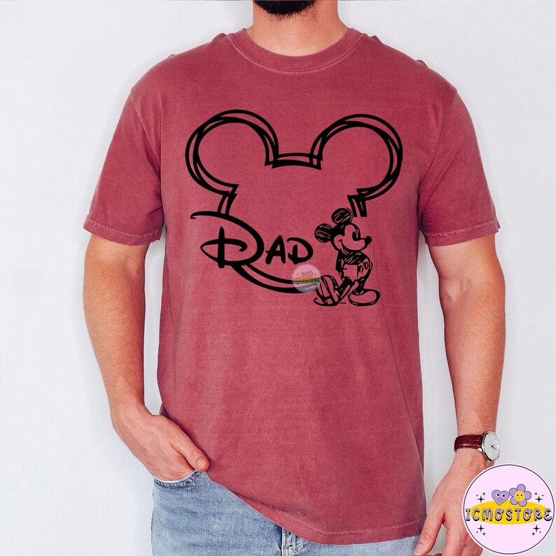 May include: A maroon t-shirt featuring a black outline of Mickey Mouse ears with the word "Dad" in a stylized font. A small Mickey Mouse graphic is on the right side. The shirt is a solid color and has short sleeves.
