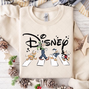 May include: Cream-colored sweatshirt featuring a Disney-themed graphic. The design depicts Disney characters Goofy, Pluto, Donald Duck, and Mickey Mouse crossing a crosswalk, with the Disney logo above. The sweatshirt is a crew neck style.