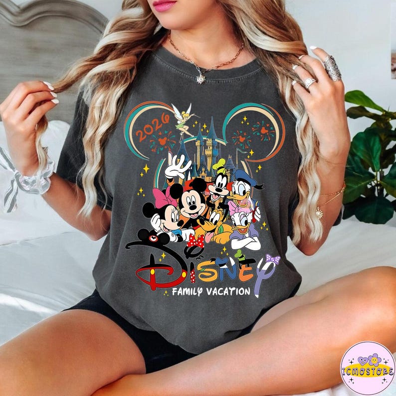 May include: Dark gray t-shirt featuring Disney characters including Mickey and Minnie Mouse, Goofy, Donald Duck, and Daisy Duck. The shirt has the text "Disney Family Vacation" and the year "2026" with a castle and fireworks design.