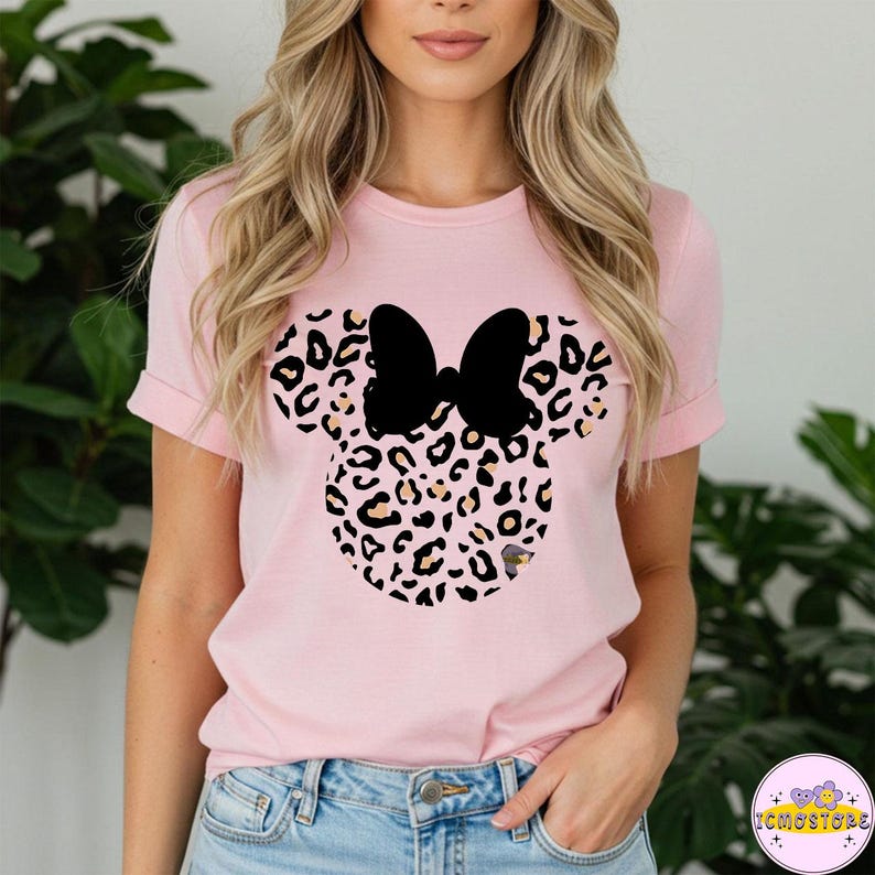 May include: A light pink t-shirt featuring a black bow and a Minnie Mouse head design with a leopard print pattern. The shirt has short sleeves and a crew neck. The design is centered on the front of the shirt.