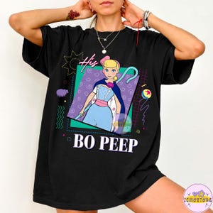 May include: Black t-shirt featuring a graphic of Bo Peep from Toy Story. The design includes the text "His" and "BO PEEP" in a retro style, with a colorful geometric background. The shirt is a casual, oversized fit.