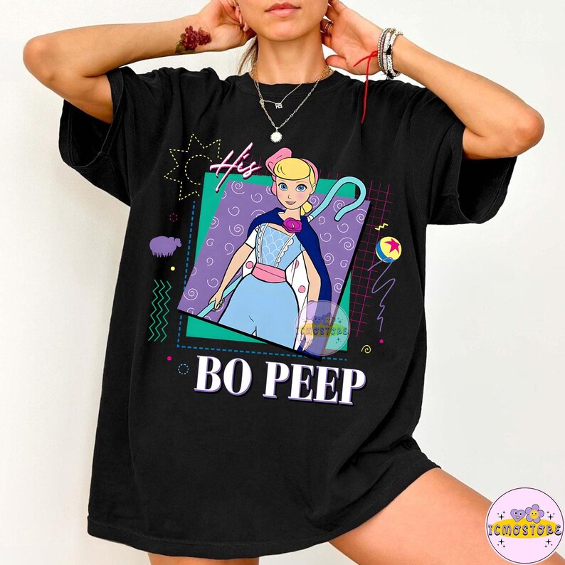 May include: Black t-shirt featuring a graphic of Bo Peep from Toy Story. The design includes the text "His" and "BO PEEP" in a retro style, with a colorful geometric background. The shirt is a casual, oversized fit.