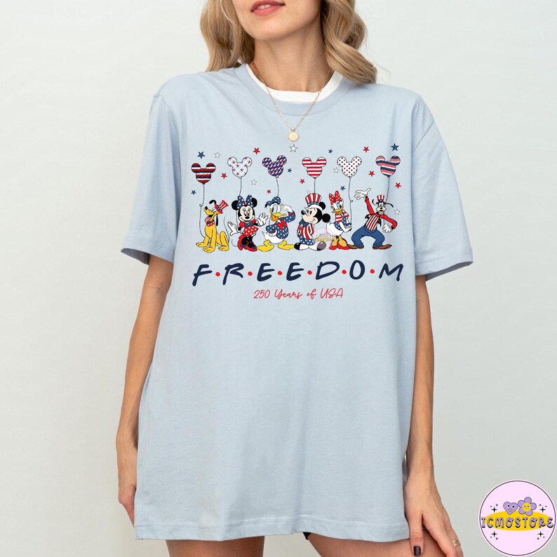 May include: Light blue t-shirt featuring a graphic of Disney characters holding patriotic balloons. The word "FREEDOM" is printed in a stylized font, with "250 Years of USA" below. The shirt is a casual style.