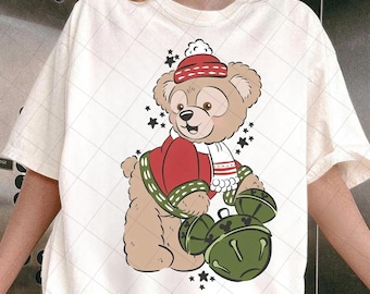 May include: Cream t-shirt with a cartoon bear in festive attire. The bear has a red hat and jacket, a white scarf, and holds green bells. Black stars are scattered around the bear. The shirt has a diamond pattern.