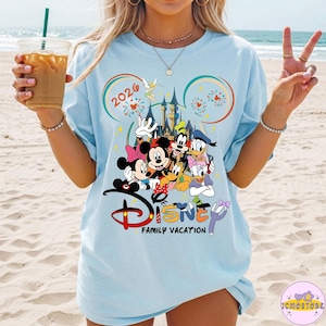 May include: Light blue t-shirt featuring a Disney family vacation design. The graphic includes Mickey and Minnie Mouse, Goofy, Donald and Daisy Duck, and a castle. The text "2024" and "Disney Family Vacation" are also visible.