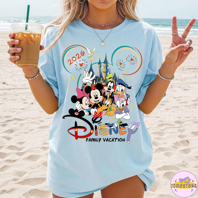 May include: Light blue t-shirt featuring a Disney family vacation design. The graphic includes Mickey and Minnie Mouse, Goofy, Donald and Daisy Duck, and a castle. The text "2024" and "Disney Family Vacation" are also visible.