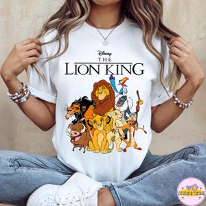 May include: White t-shirt featuring a colorful graphic of characters from Disney's The Lion King, including Simba, Mufasa, and Timon. The text "Disney The Lion King" is printed above the characters in black.