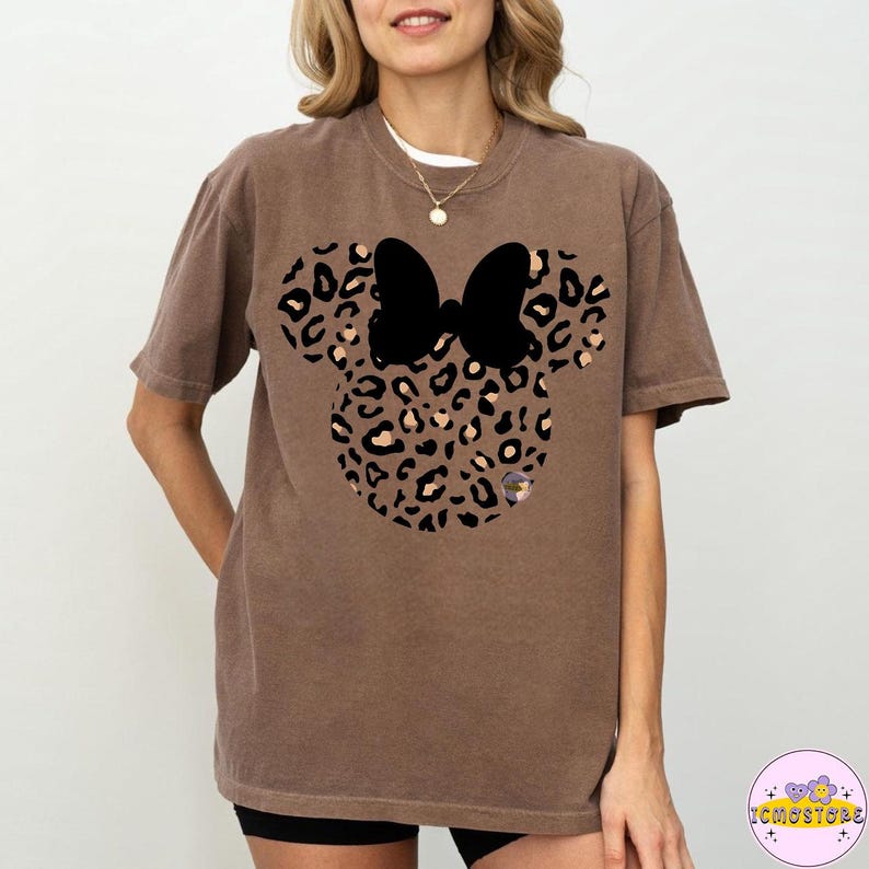 May include: Brown t-shirt featuring a Minnie Mouse design with a leopard print pattern and a black bow. The shirt is a comfortable, casual style, perfect for everyday wear. The design is centered on the front of the shirt.