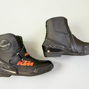 Könnte beinhalten: Paar schwarze Motorradstiefel mit orangefarbenen KTM-Logos. Die Stiefel haben ein hohes Design mit Reißverschluss und Schnallenverschluss. Sie verfügen über verstärkte Paneele und eine strukturierte Sohle.
