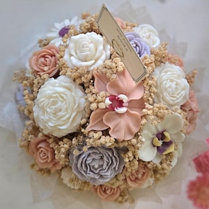 Può includere: Un bouquet di fiori artificiali con rose e orchidee bianche, color pesca e lavanda, intervallate da gipsofila essiccata. Un cartellino regalo rettangolare color beige con il testo BEST WISHES BAGRA è inserito al centro della composizione.