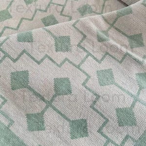 May include: A folded rug in light tones, displaying a repeating geometric pattern. The design incorporates squares and zig-zag lines in a muted green, creating a textured surface. The rug's edges are bordered with a matching green stripe.