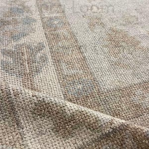Op de afbeelding: Close-up van een beige en bruin tapijt met een gedetailleerd, getextureerd patroon. Het tapijt heeft een randontwerp en subtiele blauwe accenten, wat een vintage of antieke esthetiek laat zien. Het tapijt is opgevouwen en onthult de dikte en het weefsel.
