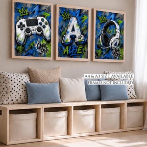 May include: Three framed wall art prints featuring graffiti-style blue, green, and black backgrounds. The prints display a white game controller, the letter A with the name ALFIE, and white gaming headphones. Text includes LEVEL UP and GAME ON.