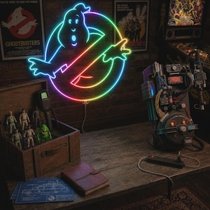 May include: A glowing neon sign featuring the Ghostbusters logo with a multi-colored gradient, positioned above a wooden crate containing small action figures, a proton pack replica, a brown leather-bound notebook, and a blue technical blueprint.