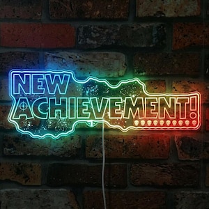 Könnte beinhalten: Ein Neon-Schild in Form eines Schlüssels mit den Worten "NEW ACHIEVEMENT!" in Blau und Weiß. Das Schild hat einen Farbverlauf, der von Blau über Grün nach Rot verläuft. Der Hintergrund ist eine Backsteinmauer.
