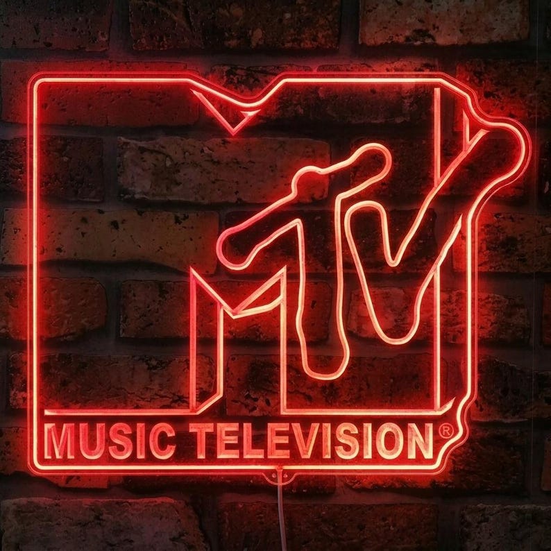 May include: A bright red neon sign shaped like the MTV logo, featuring the large block letter M and the stylised TV graphic. Below the logo, the text MUSIC TELEVISION is illuminated in the same vibrant red neon light against a dark brick wall.