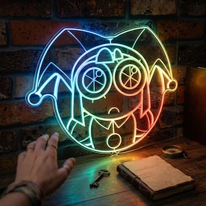 Può includere: Un'insegna al neon luminosa che raffigura un personaggio giullare stilizzato con grandi occhi circolari e un cappello a punta. La luce sfuma dal blu a sinistra all'arancione a destra, creando un vibrante effetto gradiente sulla parete.