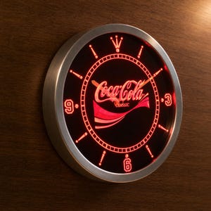 May include: A round wall clock with a brushed silver metal frame and a black face illuminated by red neon light. The centre features the Coca-Cola Classic logo in red script. The clock has red hour markers, red numerals at 3, 6, and 9, and red hands.