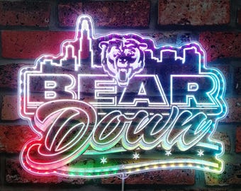 May include: A neon sign with the words "BEAR Down" in a stylized font, featuring a bear head and a city skyline silhouette. The sign has a multi-colored border with red, green, and blue hues, and is mounted on a brick wall.