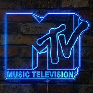 May include: A bright blue neon sign displaying the MTV logo with the text MUSIC TELEVISION underneath. The sign features a bold, outlined M and a stylized V, glowing with a vibrant blue light against a dark, textured brick wall background.