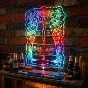 Könnte beinhalten: Ein Neonschild mit farbigem Umriss zeigt zwei Modelo-Bierflaschen, Zuckerschädel und den Text "VIVA LA TRADICIÓN". Das Schild ist an einer Backsteinmauer angebracht, mit zusätzlichen Modelo-Bierflaschen und Barzubehör.