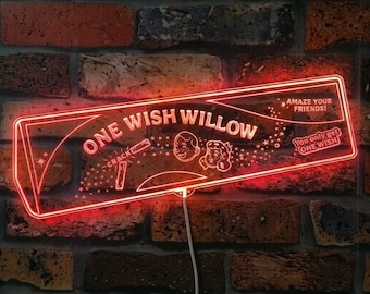 May include: A rectangular red neon sign featuring the text ONE WISH WILLOW, AMAZE YOUR FRIENDS!, and You only get ONE WISH. The design includes line art of two faces, a small stick, and star accents, with a white power cord extending downward.