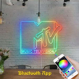 May include: A neon sign displaying the MTV Music Television logo with a multi-coloured gradient effect. The sign is mounted on a white brick wall and is controlled by a smartphone app showing a colour wheel interface for adjusting the light settings.