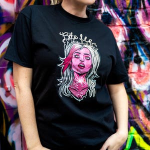 May include: Black t-shirt with a graphic of a woman with pink hair and the text "Bite Me". The illustration has a gothic aesthetic. The background is a colourful graffiti wall.