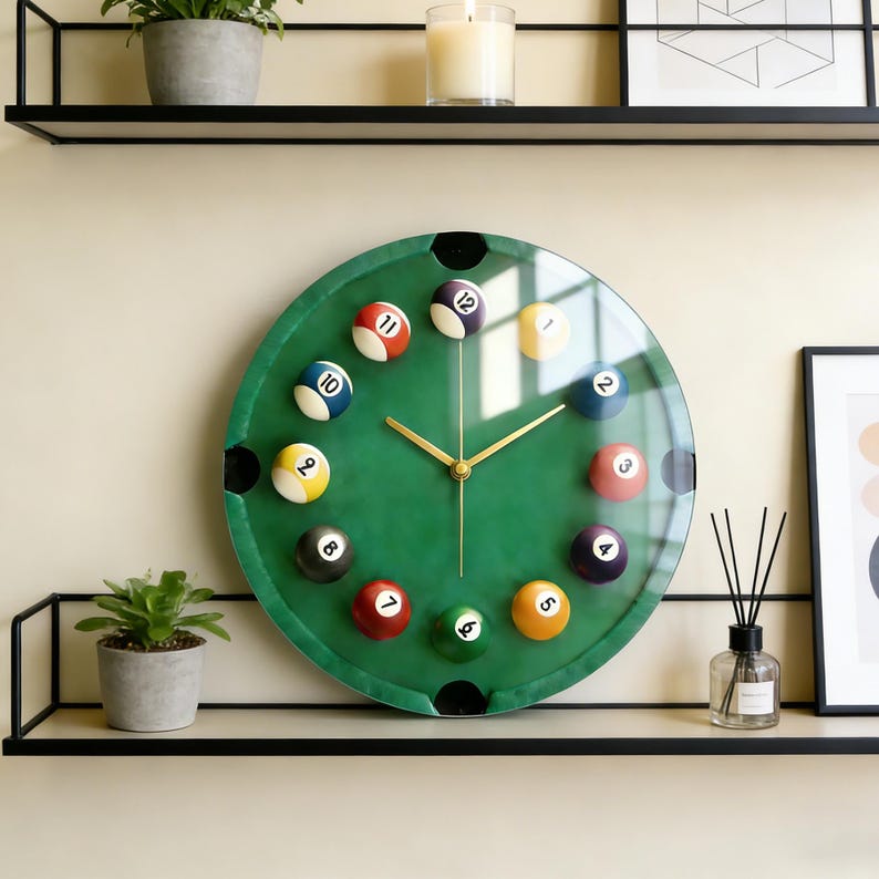 May include: A round wall clock with a green face designed to resemble a pool table. The clock features billiard balls marking the hours, gold-colored hands, and black corner pockets. The clock is displayed on a shelf.