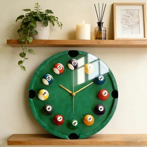May include: A green billiard table-themed wall clock with gold hands and numbered billiard balls marking the hours. The clock has a glass cover and is mounted on a wall. The clock is a decorative item.