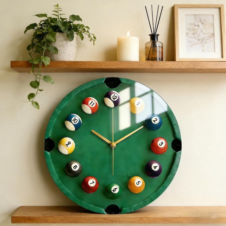 May include: A green billiard table-themed wall clock with gold hands and numbered billiard balls marking the hours. The clock has a glass cover and is mounted on a wall. The clock is a decorative item.