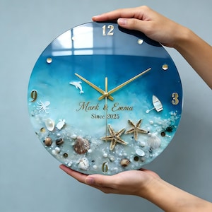 May include: A round wall clock with a beach theme. The clock face features a blue and white ocean design with seashells, starfish, and a small boat. The clock hands and numerals are gold. The text "Mark & Emma Since 2025" is displayed.