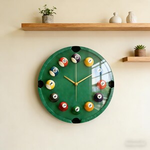 May include: A round wall clock with a green face designed to resemble a pool table. The clock features billiard balls in place of numbers and gold-colored hands. The clock is mounted on a light-colored wall.