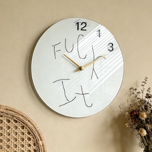 May include: A round, white wall clock with black handwritten text that reads "FUCK IT". The clock has gold-colored hands and black numbers at the 12, 1, and 3 o'clock positions. The clock is mounted on a beige wall.