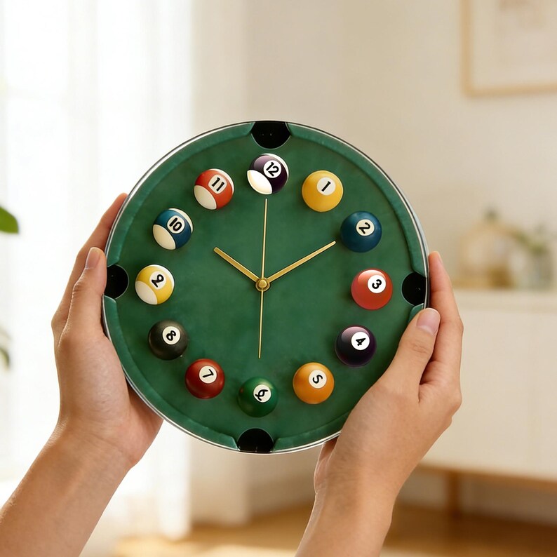 May include: A round wall clock with a green felt face, resembling a pool table. The clock features billiard balls marking the hours, with gold-colored hands. The clock is held by two hands.