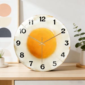 May include: A round wall clock with a fried egg design. The clock face features a yellow yolk surrounded by a white egg white, with black numerals marking the hours. The clock hands are gold-colored.