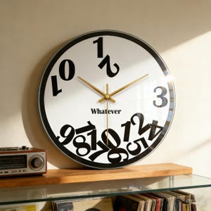 May include: A round wall clock with a white face and black numbers in a unique, scattered arrangement. The clock hands are gold, and the word "Whatever" is printed below the center. The clock is displayed on a wooden shelf.