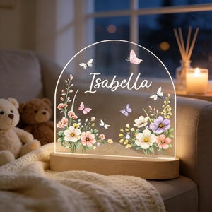 May include: A personalized night light with the name "Isabella" in white script. The clear acrylic arch features floral and butterfly designs in pastel colors, set on a wooden base. The lamp is illuminated, creating a soft glow.
