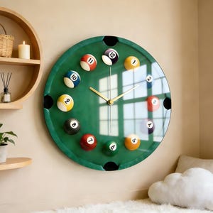 May include: A round wall clock with a green felt-like surface, resembling a pool table. The clock features billiard balls as hour markers and gold-colored hands. The clock is mounted on a beige wall.
