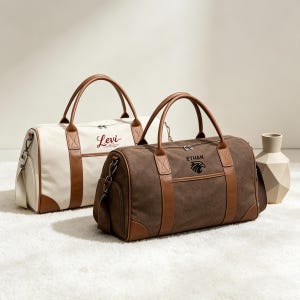 May include: Two canvas duffel bags with brown leather accents and handles. One bag is off-white with "Levi" in red script, the other is brown with "Ethan" and an eagle graphic in black. Both have side pockets.