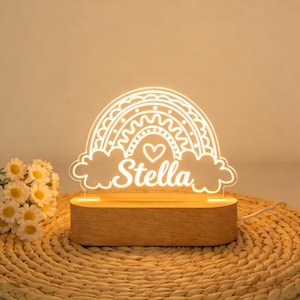 May include: A glowing LED rainbow night light with the name "Stella" in a script font. The rainbow design includes a heart and cloud accents. The light sits on a rectangular wooden base and is plugged in.