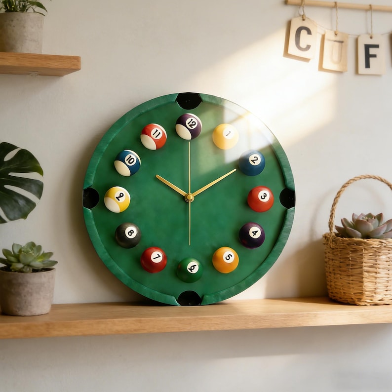 May include: A round wall clock with a green surface designed to resemble a pool table. The clock features gold-colored hands and is surrounded by billiard balls in various colors, each with a number from 1 to 12. The clock is displayed on a wooden shelf.