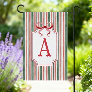 May include: A garden flag with a red letter "A" inside a white frame, adorned with a red bow. The flag features vertical stripes in red and green, creating a festive and personalized outdoor decoration.
