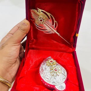May include: A silver-toned feather brooch and a matching oval-shaped pendant, both adorned with red and clear gemstones, are displayed in a red velvet box. The brooch has a detailed feather design. The box is closed with a gold-colored clasp.