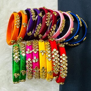 May include: A collection of colorful bangles in various shades, including orange, purple, red, pink, and blue. Each bangle is adorned with gold-colored embellishments and small, clear stones. The bangles are arranged on a white and black surface.
